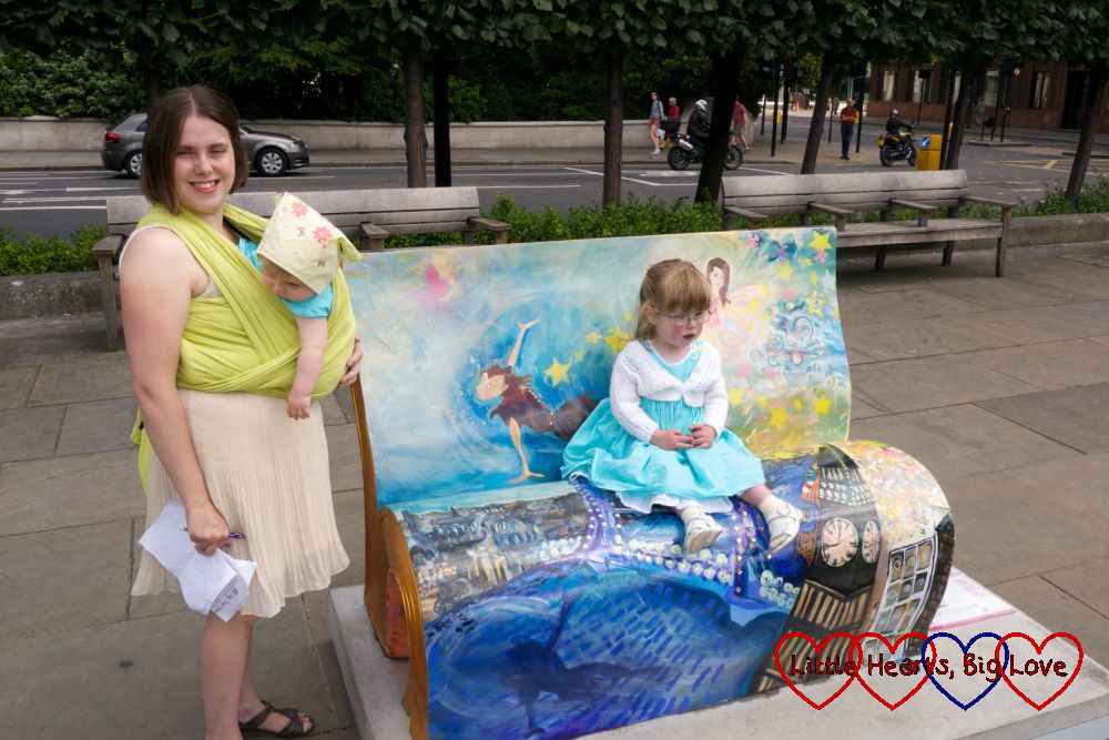 A little Jessica sitting on a Peter Pan-themed book bench with me standing next to the bench with baby Sophie in a wrap