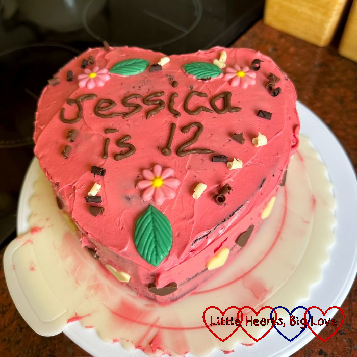 A heart-shaped cake with dark pink icing, pink chocolate flowers and milk and white chocolate curls and the words "Jessica is 12" piped in chocolate writing.