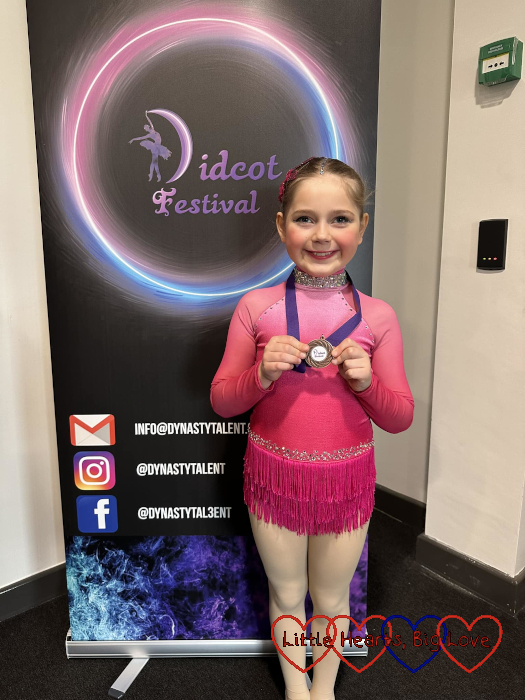 Sophie in a pink leotard with a fringed skirt, standing in front of a 'Didcot festival' board, holding a bronze medal