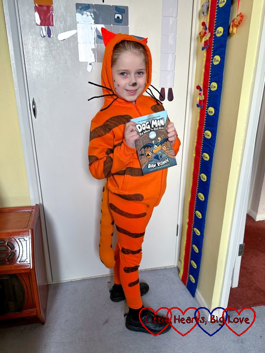 Sophie wearing an orange striped hoody and leggings with an orange striped tail and holding a Dogman book.