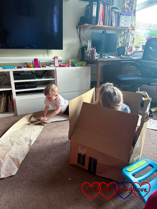Sophie sitting in a cardboard box while Thomas uses the paper packaging as a track for a toy car